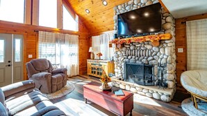 A cozy log cabin interior with a stone fireplace, a television mounted above it, and a comfortable seating area with a sofa and armchairs. - Panoramic Point - 91 Panoramic Drive (Dryfork)
