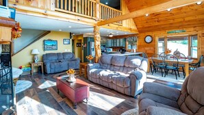 A spacious living area with a high ceiling, wooden beams, a dining area with a large table and chairs, and a kitchen in the background. - Panoramic Point - 91 Panoramic Drive (Dryfork)