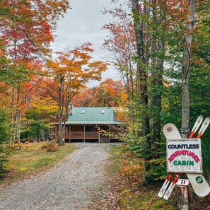 Una cabaña con la leyenda 'Countless Adventures Cabin', con un camino de grava que la lleva a ella, rodeada de árboles de colores otoñales.