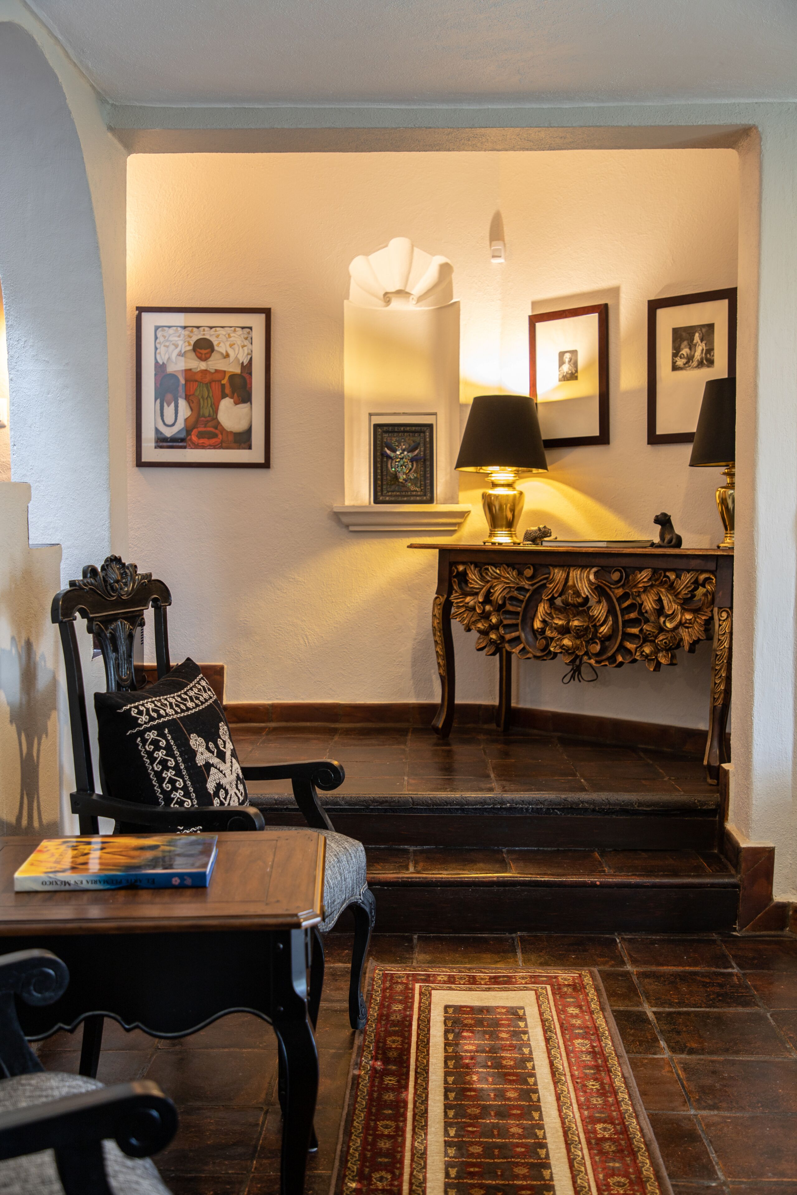A room with a wooden chair, a table with a book, a patterned rug, a console table with lamps, and framed pictures on the wall.