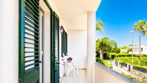 A balcony with a white table and chairs, green shutters, and a view of greenery and a building.