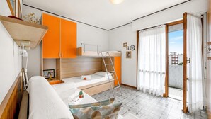 Een compacte kamer met een stapelbed, een slaapbank, een klein tafeltje en een balkon met gordijnen.