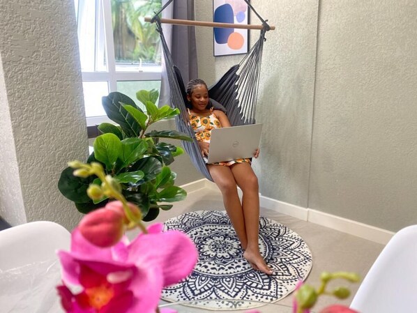 Une personne assise dans un fauteuil suspendu en forme de hamac, un ordinateur portable sur les genoux, entourée de plantes d’intérieur et posée sur un tapis au motif décoratif.