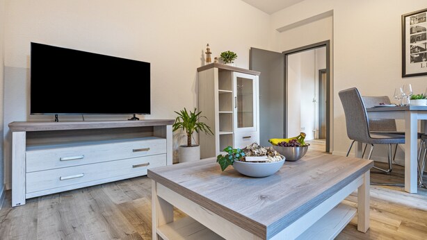 Una sala de estar moderna con un televisor de pantalla plana, una zona de comedor con mesa y sillas, y un mueble blanco con puertas de cristal.