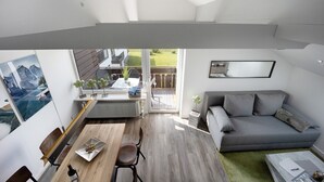 A modern living room with a grey sofa, a dining table with chairs, a view of the outdoors, and a large window. - Studio Apartment: Loft with Lake View (Petersthal)