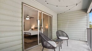 Un balcon avec deux fauteuils en osier, un ventilateur de plafond et une porte-fenêtre coulissante donnant sur une chambre à coucher équipée d’un lit, d’une table de chevet et d’une lampe.