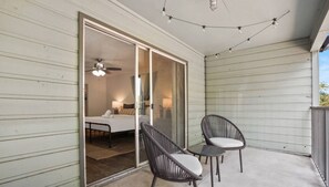 Un balcón con dos sillas de mimbre, un ventilador de techo y un cerramiento de vidrio corredera que da a un dormitorio con cama, mesita de noche y lámpara.