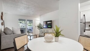 Un salon moderne avec une table ronde blanche, deux plantes en pot, un canapé gris et un téléviseur à écran plat diffusant du contenu Hulu.