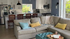 A modern living room with a grey sofa, teal ottoman, and wooden chairs. A kitchen with white cabinets and a large window is visible in the background. - Peaceful retreat with fully stocked kitchen. (Redlands)