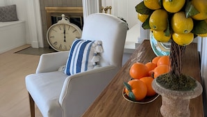 A living room with a white armchair, a wooden side table with oranges and a decorative plant, a fireplace, and a large wall clock. - Peaceful retreat with fully stocked kitchen. (Redlands)