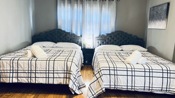 A room with two beds, each with a headboard and a white and black checkered bedspread. There is a window with curtains, a wooden floor, and a painting on the wall. - Luxury 2 bedroom 🛌 Next to UBS Arena🏟 & JFK✈️ (Valley Stream)