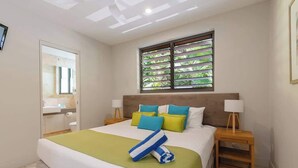 A hotel room with a bed, a window with blinds, a bedside table with a lamp, and a bathroom with a mirror and sink. - Villa On The Hill —Spacious Tropical Panorama (Port Douglas)