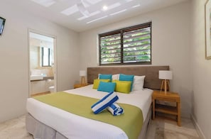 Une chambre d’hôtel comprenant un lit, une fenêtre avec des stores, une table de chevet avec une lampe et une salle de bain avec un miroir et un lavabo.