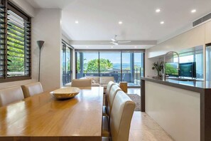 A modern dining area with a wooden table, leather seating, and large windows offering a view of greenery and hills.