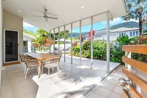 Un soggiorno open space con tavolo e sedie, una ventola a soffitto e vista su un giardino rigoglioso e case.