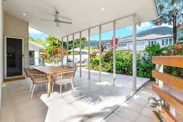 Un espace de vie à aire ouverte comprenant une table et des chaises, un ventilateur de plafond, et donnant sur un jardin luxuriant et des maisons.