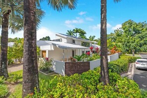 Une maison moderne à deux étages, à l’extérieur blanc, entourée d’une végétation luxuriante et de palmiers.