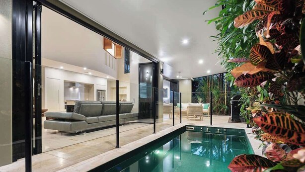 Ein moderner Poolbereich im Innenbereich mit Glasbarriere, grauem Sofa und tropischen Pflanzen.