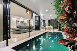Ein moderner Poolbereich im Innenbereich mit Glasbarriere, grauem Sofa und tropischen Pflanzen.