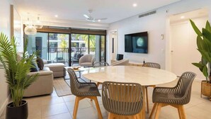 A modern living room with a round dining table, comfortable seating, a flat-screen TV, and large windows. - Sunny Swim-out Serenity with Direct Beach Access (Port Douglas)