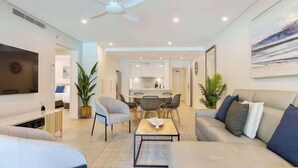 A modern living room with a sofa, coffee table, and chairs, a dining area with a table and chairs, and a kitchen in the background. - Sunny Swim-out Serenity with Direct Beach Access (Port Douglas)