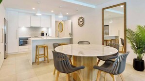 A modern kitchen with white cabinets, a round dining table with chairs, a large mirror, and a clock on the wall. - Sunny Swim-out Serenity with Direct Beach Access (Port Douglas)