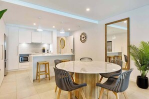 A modern kitchen with white cabinets, a round dining table with chairs, a large mirror, and a clock on the wall. - Sunny Swim-out Serenity with Direct Beach Access (Port Douglas)