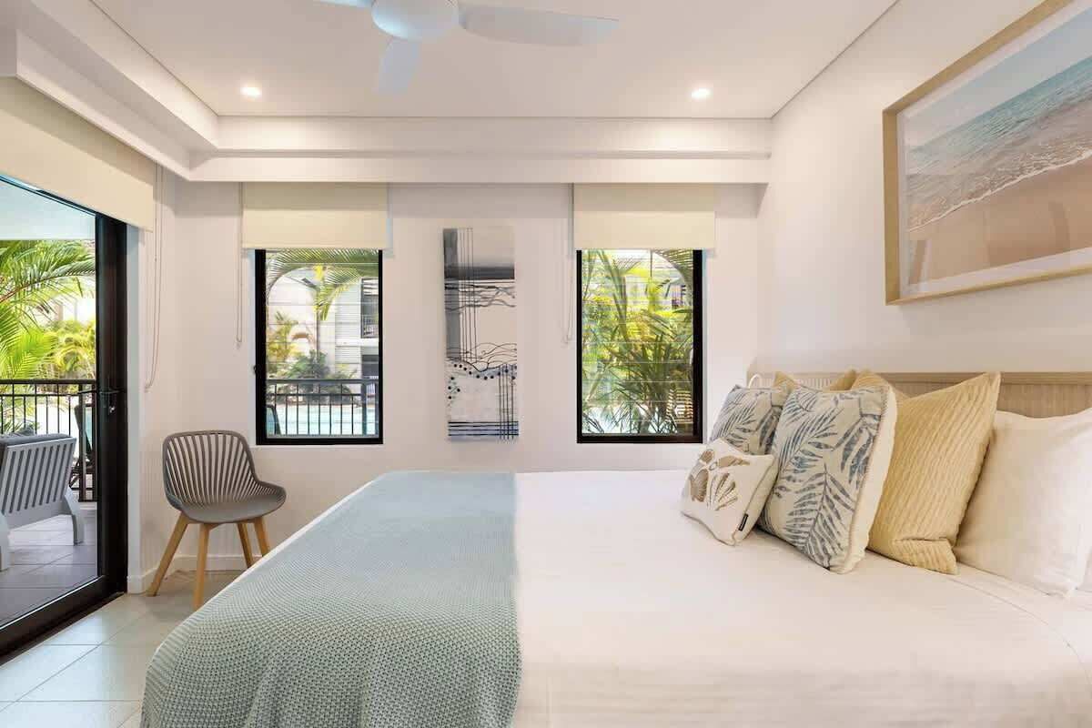 Une chambre dotée d’un grand lit, d’une chaise, d’un ventilateur de plafond et offrant une vue sur l’extérieur grâce à de grandes fenêtres.