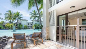 Un espace aménagé au bord de la piscine, avec deux chaises longues en bois, un mur en pierre et une vue sur un immeuble résidentiel doté d’une piscine.
