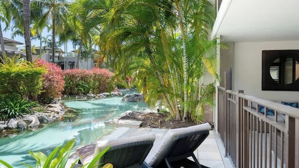 Un área de piscina con sillones de descanso, palmeras y una pequeña cascada.
