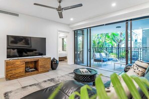 A modern living room with a flat-screen TV, a wooden entertainment unit, a black leather sofa, a coffee table, and a sliding glass door leading to an outdoor balcony with a view of greenery. - Resort-style Swim Out: A Poolside Oasis (Port Douglas)