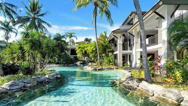 A resort with a clear pool, palm trees, and a two-story building.