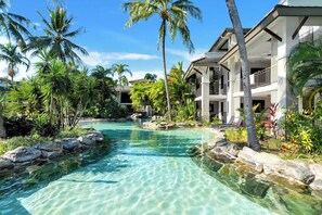 Ein Resort mit klarem Pool, Palmen und einem zweigeschossigen Gebäude.