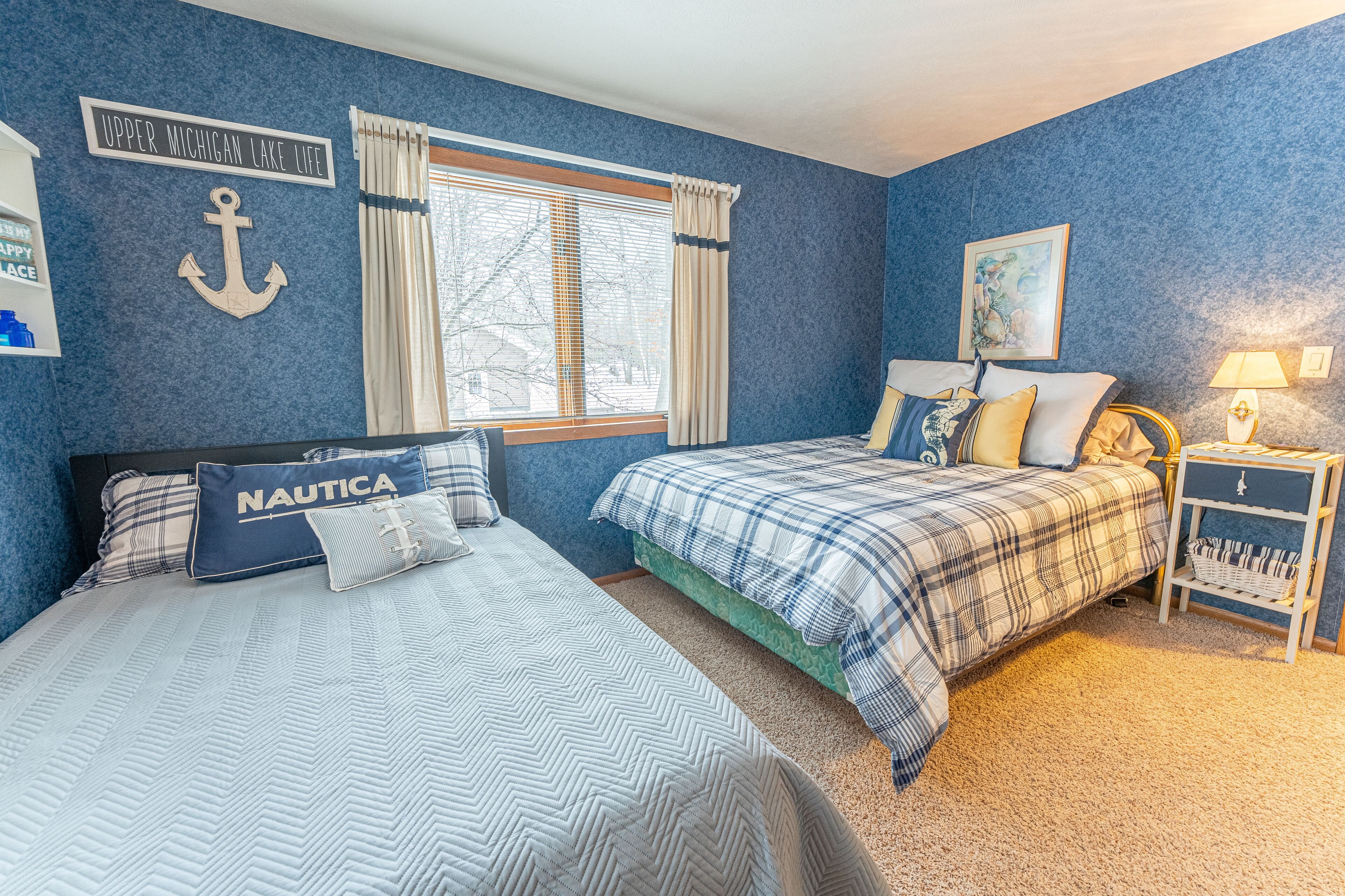 A bedroom with two beds, nautical-themed decor, a window with curtains, and a nightstand with a lamp.