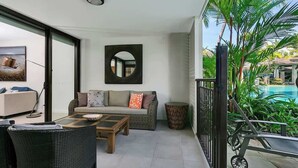 A balcony with a sofa, coffee table, wicker chairs, a round mirror, and a view of a swimming pool.