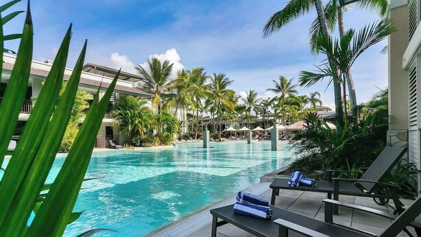 A swimming pool with lounge chairs and umbrellas, surrounded by palm trees and a building with balconies.