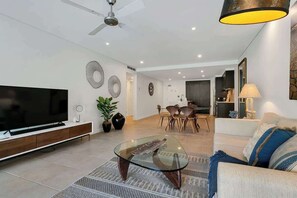Ein modernes Wohnzimmer mit Flachbildfernseher, Glastisch, Sofa, Essbereich und Deckenventilator.