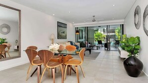 A modern dining area with a glass table and wooden chairs, a large mirror, and a view into the living room.
