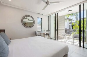 A bedroom with a bed, a ceiling fan, a round mirror, a small table with chairs, and a sliding glass door leading to an outdoor area with a balcony.