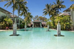 Piscina de resort con palmeras, una zona cubierta y un edificio al fondo.