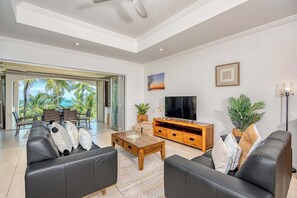 A modern living room with a TV, leather sofas, a wooden coffee table, and a large window with a view of palm trees.