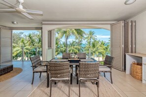 A dining area with a table and chairs, a ceiling fan, and a view of palm trees and the ocean.