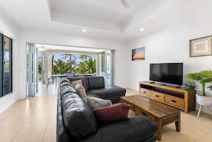 A modern living room with a leather sofa, a wooden coffee table, a flat-screen TV, and a balcony with a view of palm trees. - Verde at Island Views—Palm Cove Waterfront Bliss (Palm Cove)