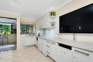 A modern kitchen with white cabinetry, a large black TV, and a window with blinds.
