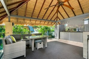 Un patio exterior con mobiliario de mimbre, ventilador de techo y una cocina con gabinetes grises.