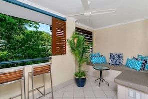A balcony with wooden chairs, a potted plant, a small table, and a sofa with patterned cushions.