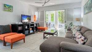 Uma sala de estar com sofá, pufê, televisão, estante de livros e mesa de centro de vidro.