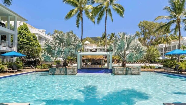 Piscina com espreguiçadeiras e guarda-sóis, rodeada por palmeiras e um edifício com varandas.