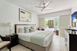 A bedroom with a bed, bedside tables, a ceiling fan, a window with blinds, a TV, and a bathroom visible through an open door.