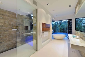 Un bagno moderno con box doccia in vetro, una grande vasca da bagno indipendente e un'ampia finestra con vista sulla vegetazione tropicale.